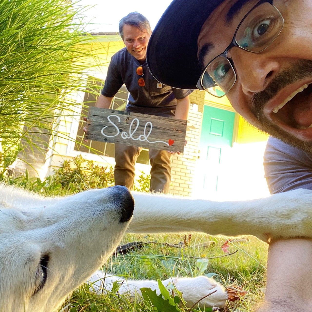 Tim taking a low angle selfie with a client's white dog while the seller holds a wooden Sold sign by a green front door
