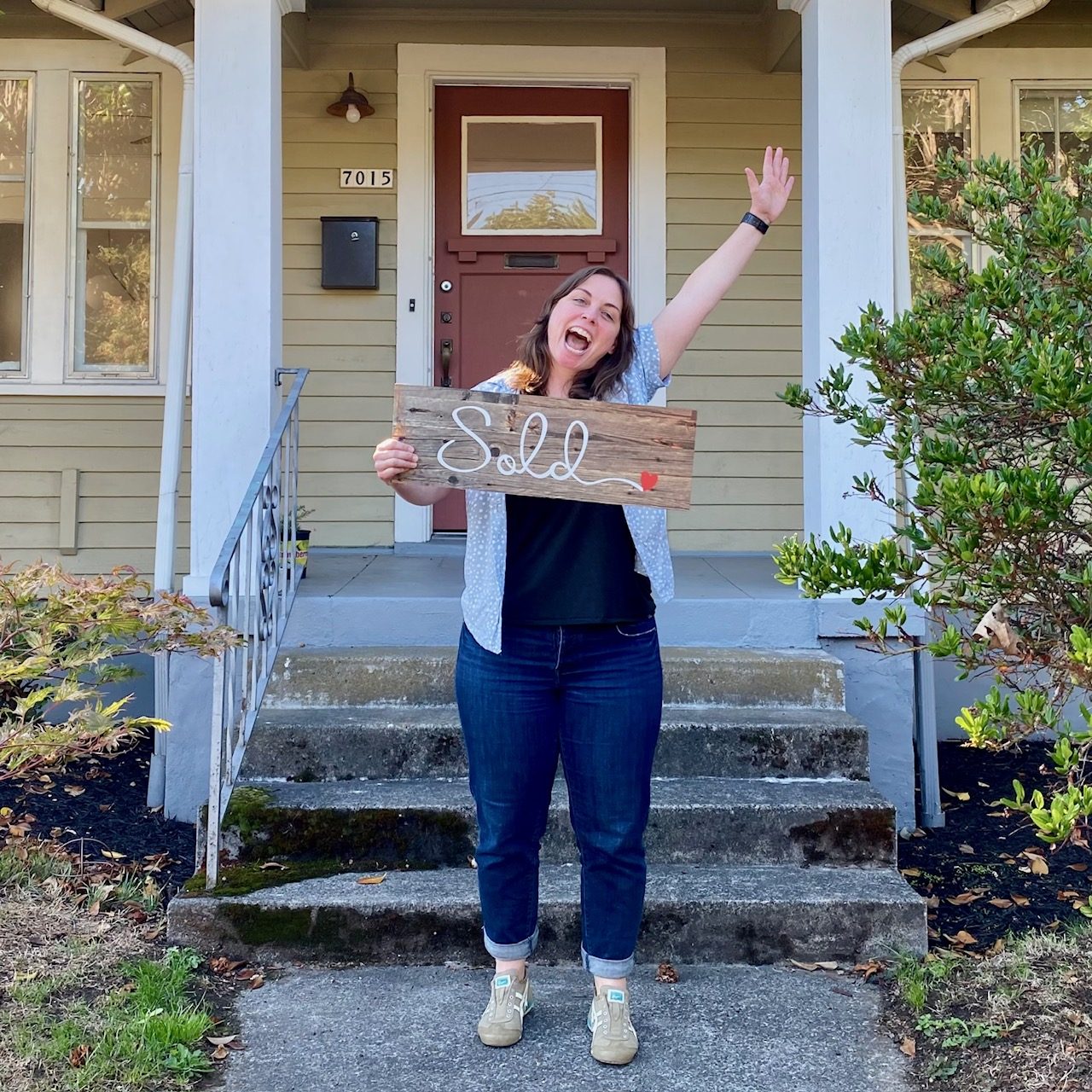 A seller cheering with one arm raised while holding a wooden Sold sign on the porch steps of her olive green Portland bungalow