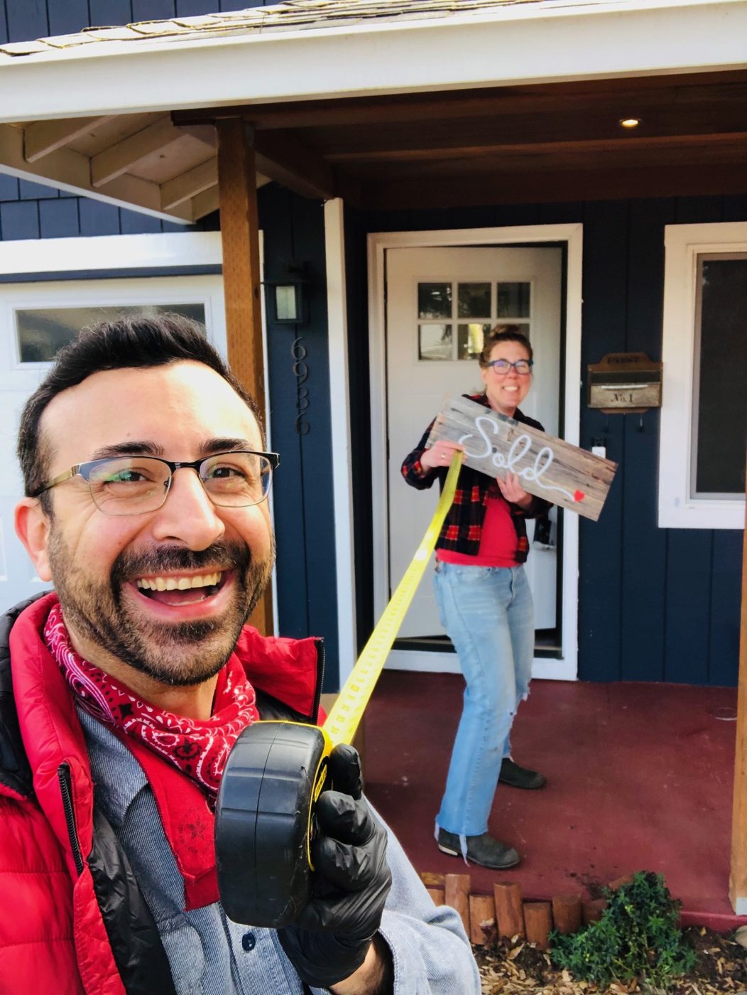 Tim holding out a tape measure in a selfie while a seller laughs behind him with a wooden Sold sign at the door of her navy blue house
