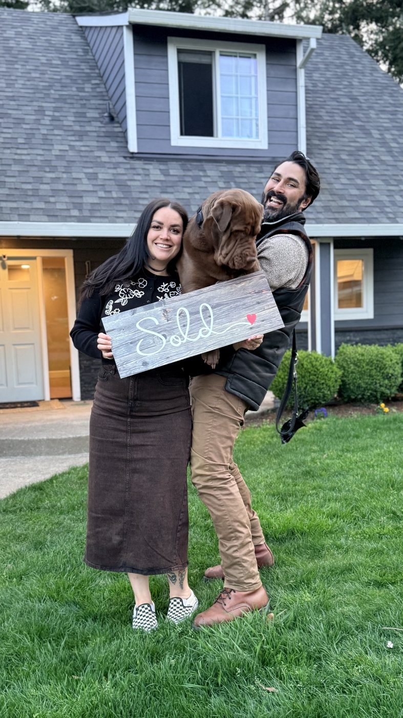 A couple lifting their large brown dog while holding a wooden Sold sign on the lawn of their new gray home
