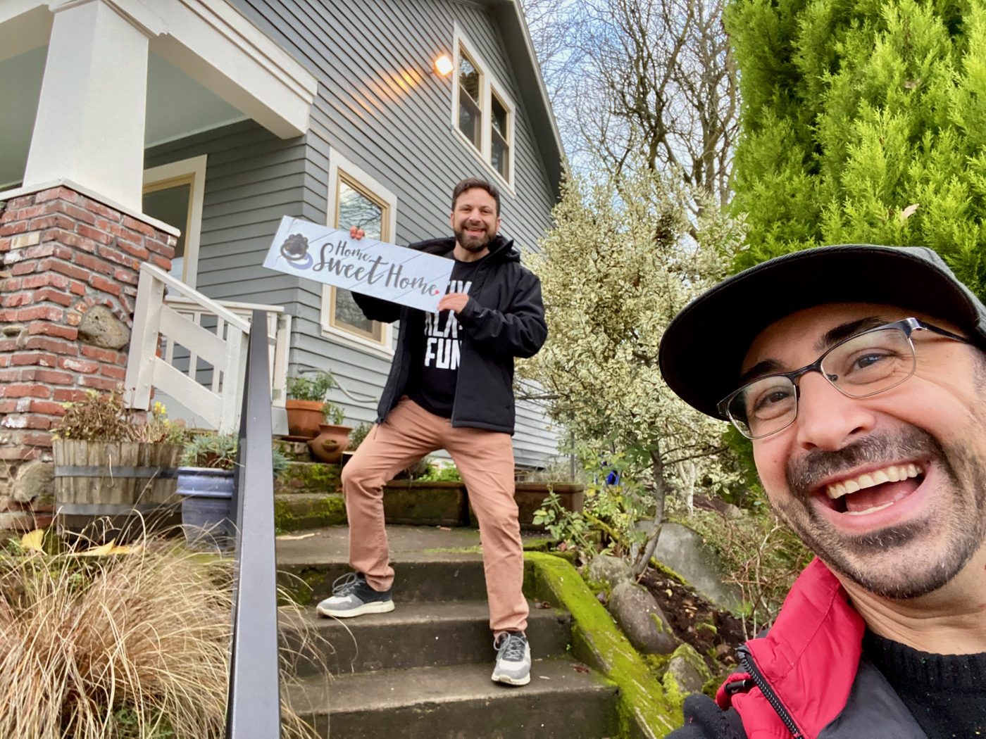 Tim smiling in a selfie while his client holds a Home Sweet Home sign on the mossy front steps of a gray Portland craftsman
