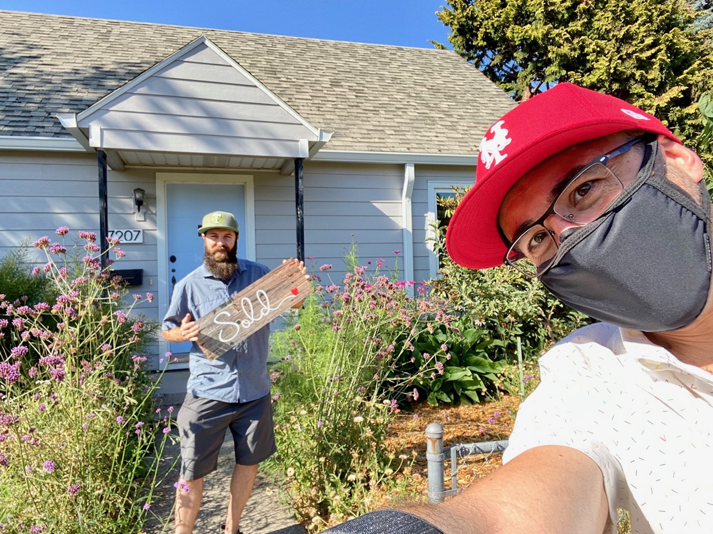 Tim in a red cap and face mask taking a selfie while his client holds a weathered Sold sign among tall purple flowers outside a pale blue cottage