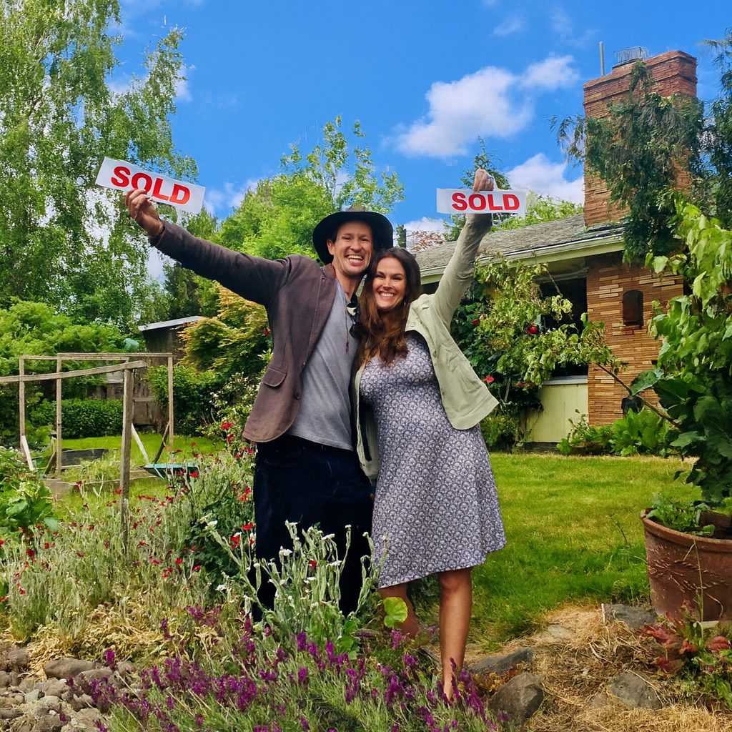 A couple raising two red Sold signs in the flower filled garden of their brick Portland home on a sunny day