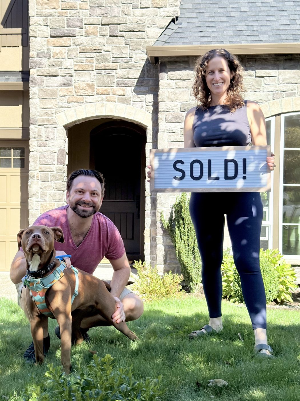 A couple with their brown dog holding a letterboard reading Sold on the front lawn of their stone facade house