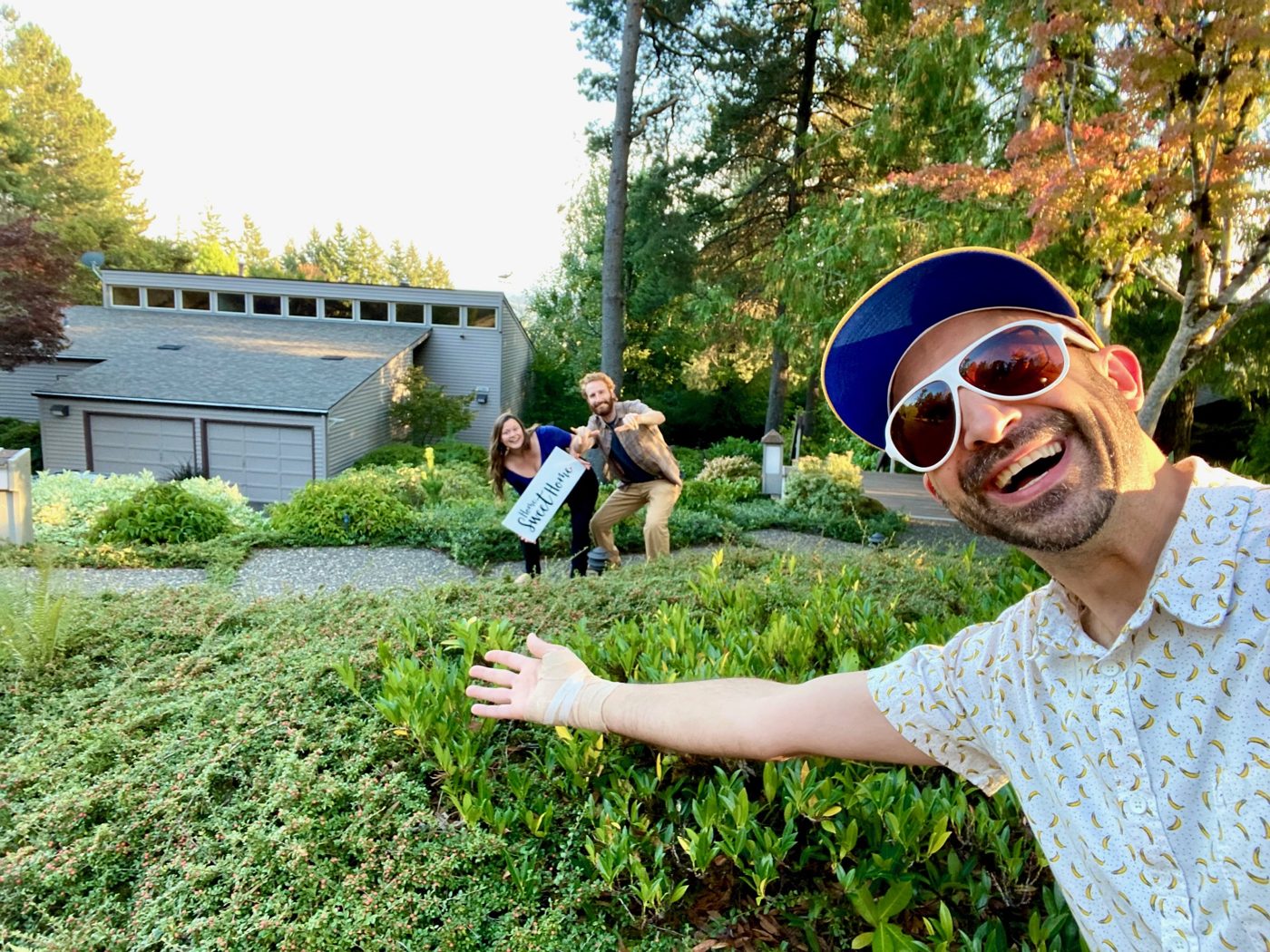 Tim leaning into a wide angle selfie in sunglasses and a ball cap while two buyers pose with a Home Sweet Home sign outside their midcentury home