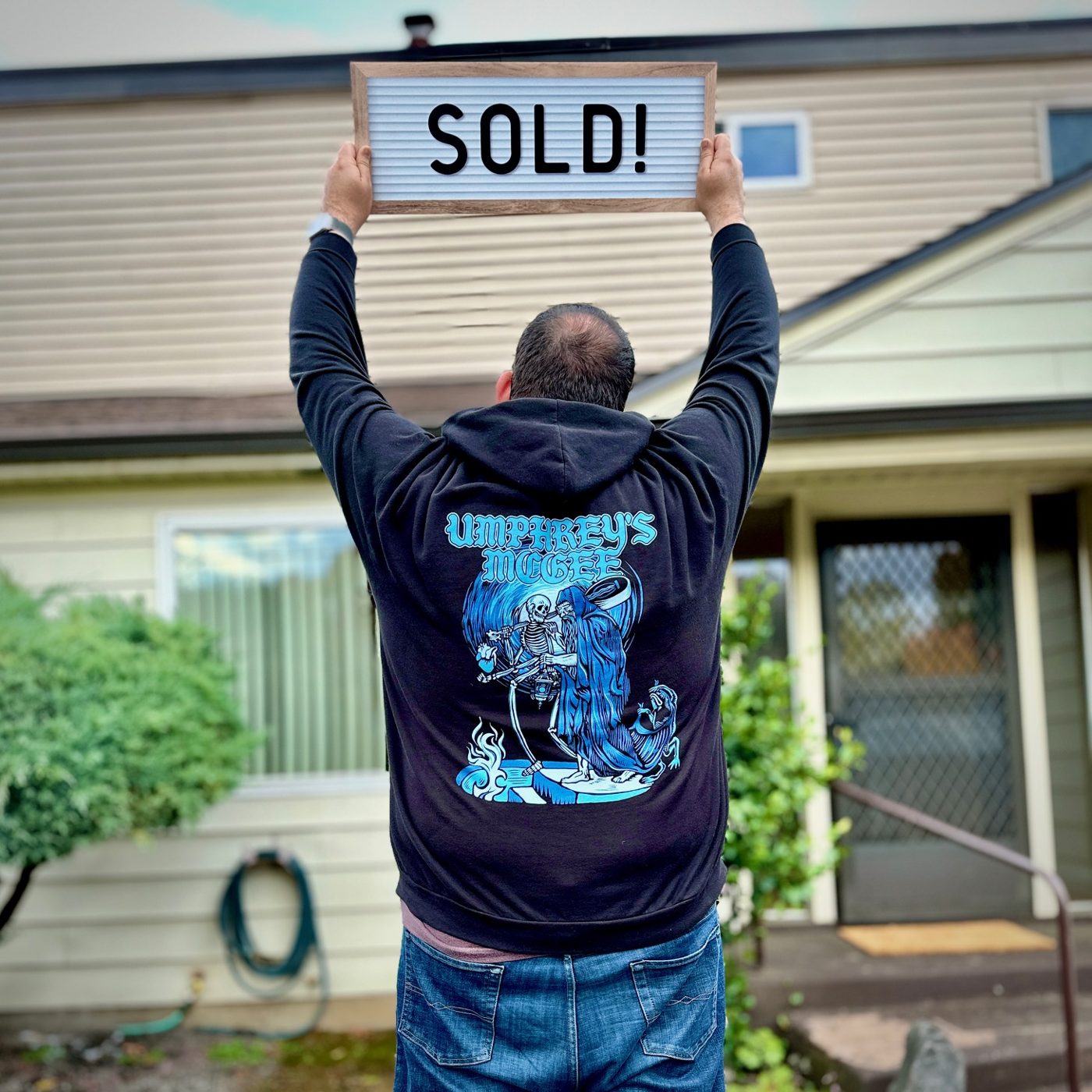 A seller seen from behind holding a letterboard reading Sold above his head in front of his home
