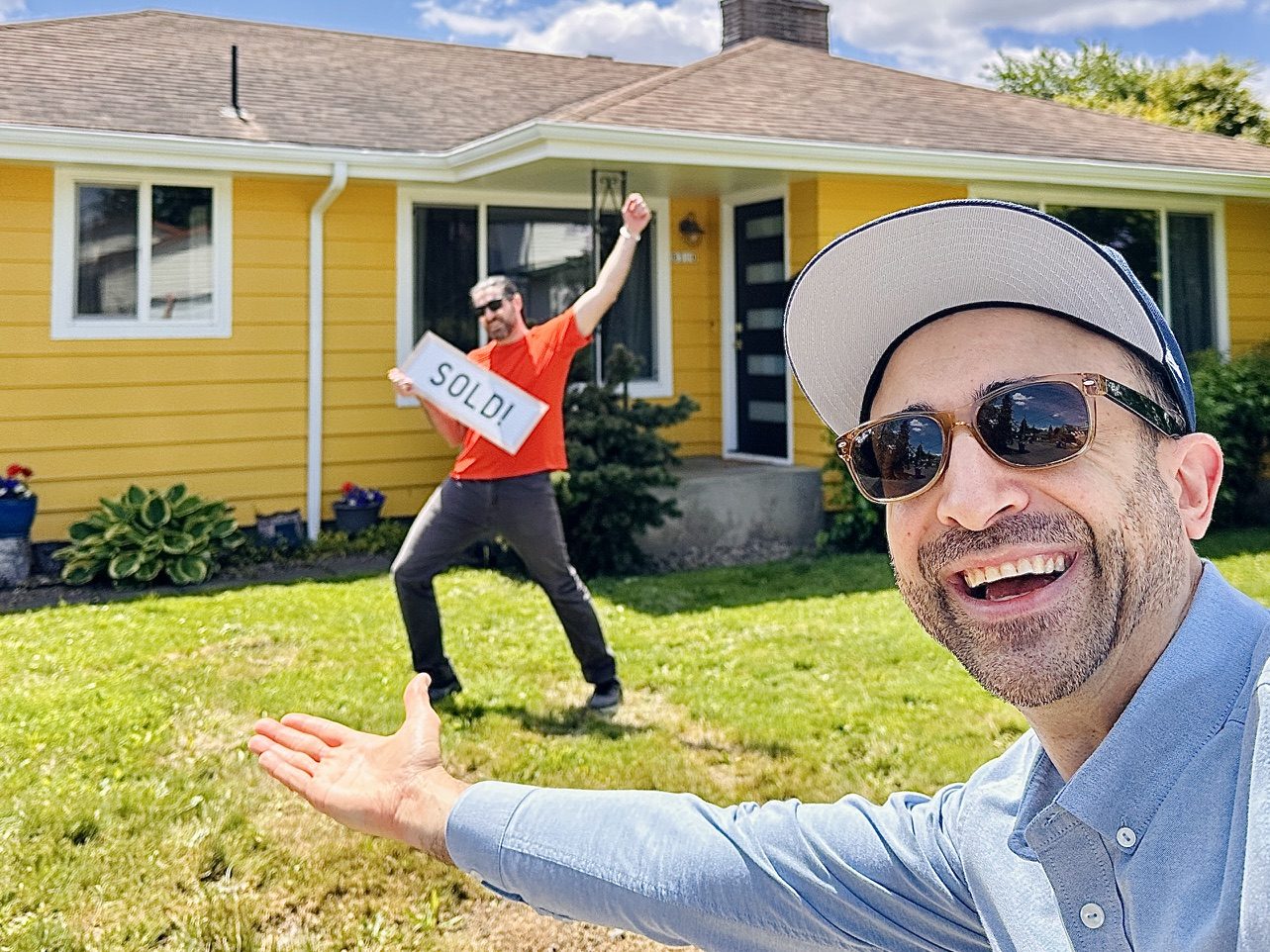 Tim in a ball cap grinning in a selfie while his client celebrates with a Sold sign in front of a bright yellow ranch house
