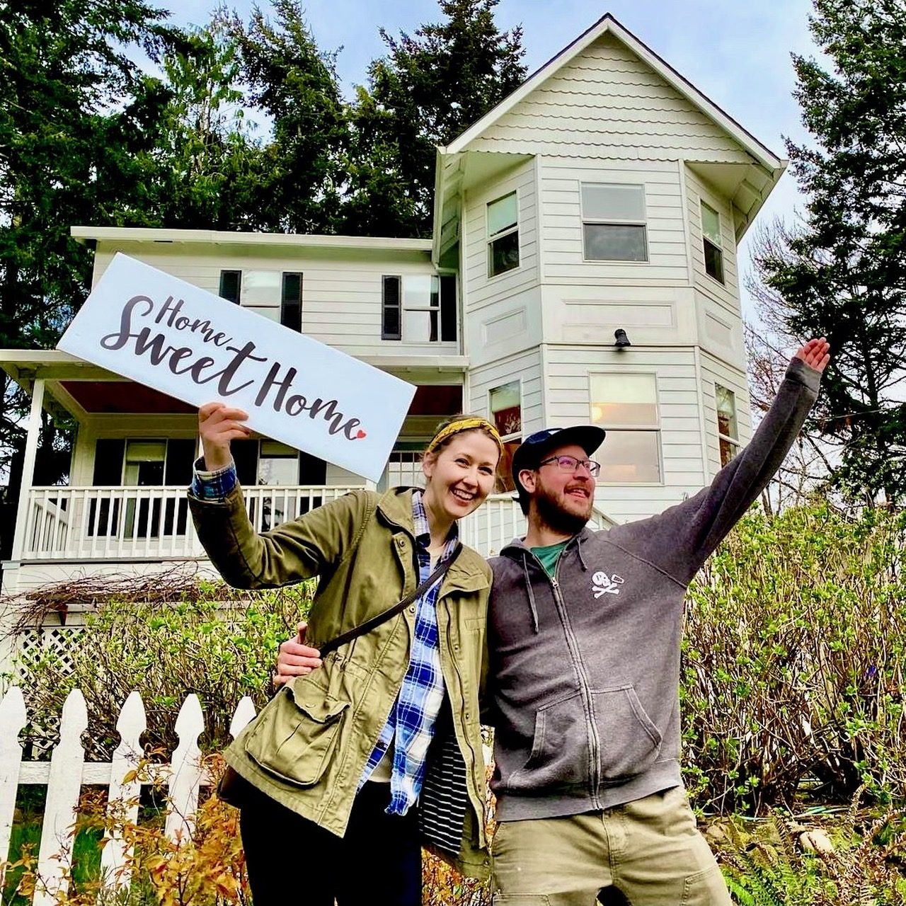 Buyers celebrating with a Home Sweet Home sign in front of a tall white Victorian house behind a picket fence