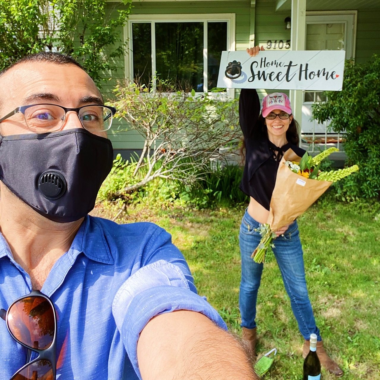 Tim in a face mask taking a selfie while his buyer holds a Home Sweet Home sign overhead with a bouquet of flowers outside her sage green house