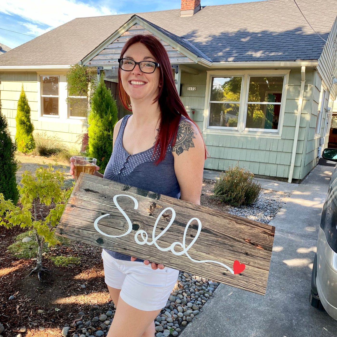 A seller with a drink in hand holding a large wooden Sold sign in the driveway of her mint green cottage