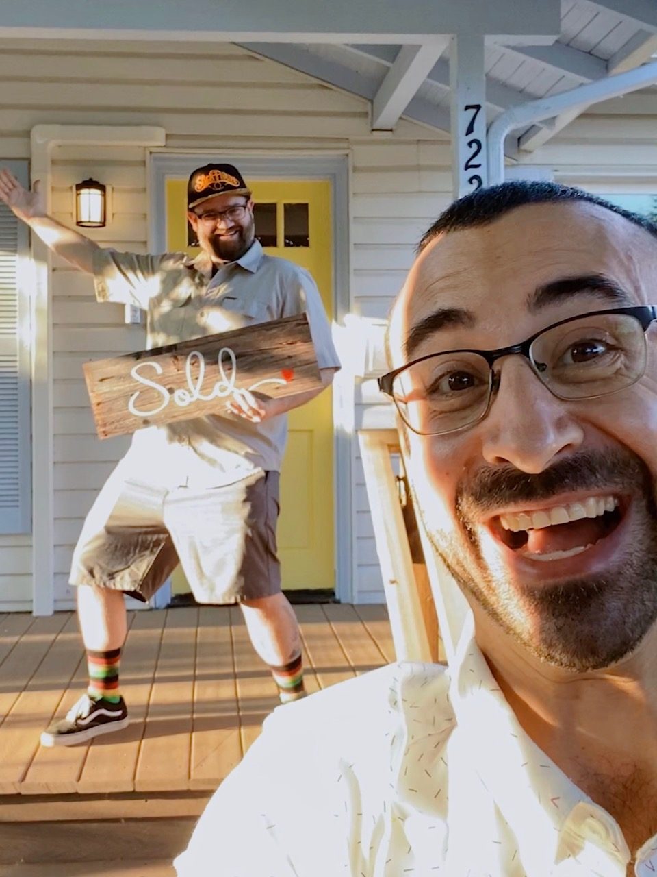 Tim laughing in a close selfie while his client strikes a triumphant pose with a reclaimed wood Sold sign on a sunlit white porch with a yellow door