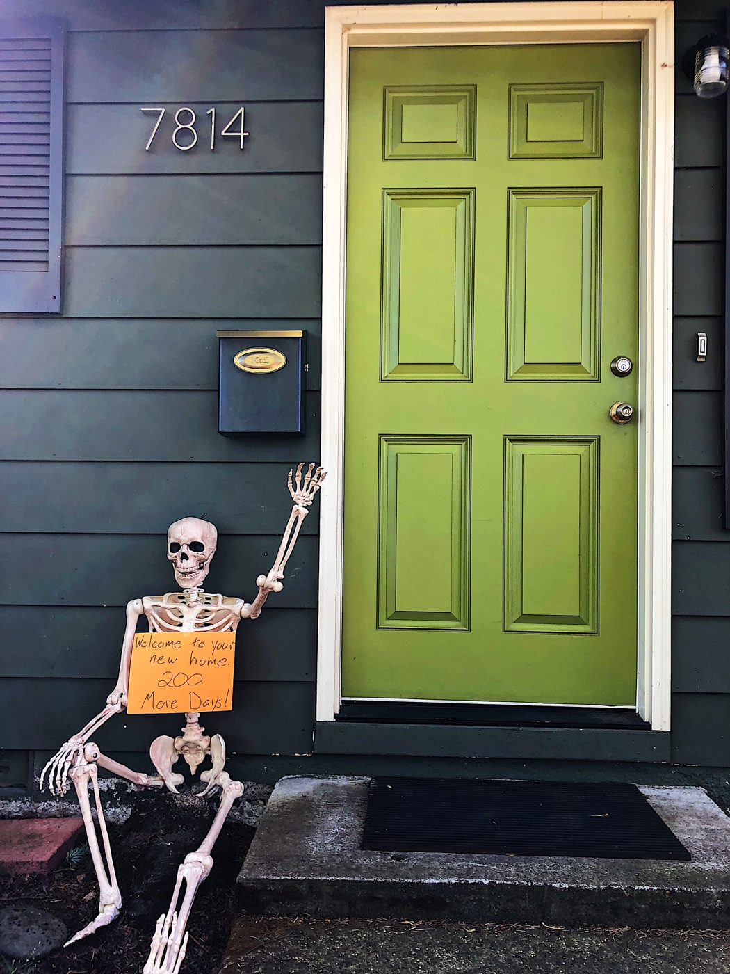 A Halloween skeleton propped by a chartreuse front door waving and holding a handwritten sign welcoming the new owners home