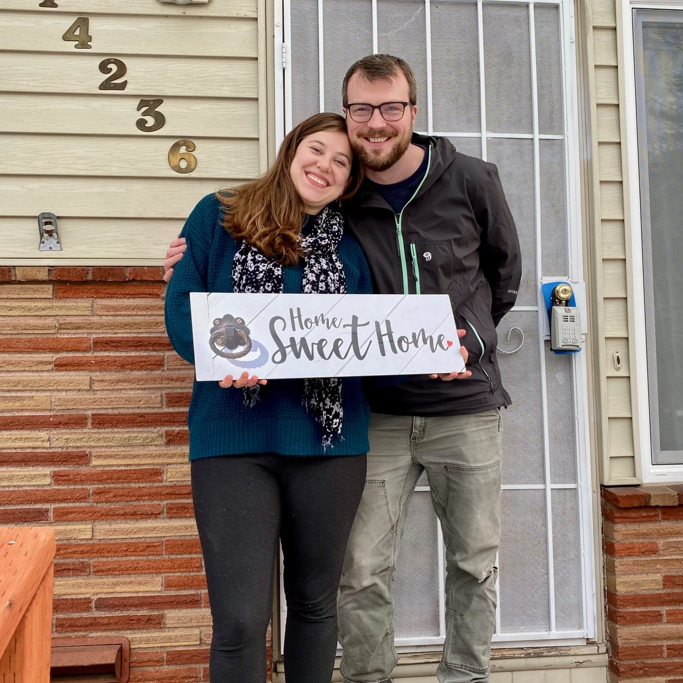 A couple standing arm in arm on their front step holding a Home Sweet Home sign beside a brick and siding entry